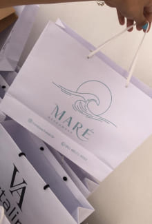 Sacola de papel na cor branca., com a logomarca da Maré (o desenho de uma onda e o nome maré) em detalhes verdes.