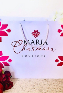 Sacola de papel colorida, com a logomarca da Maria Charmosa (a logomarca consiste em uma frase "Maria Charmosa" e um desenho de uma flor em cima dessa frase). Nos quatro cantos da sacola, tem um desenho exibindo a metade de uma flor na cor rosa.