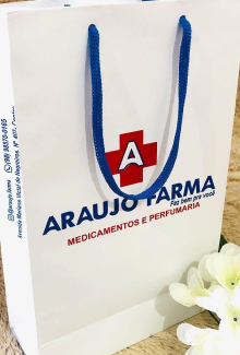 Sacola de papel colorida, com a logomarca Araujo Farma (a logomarca consiste em uma frase "Araujo Farma", com um sinal de mais na cor vermelha, e dentro desse sinal, tem a letra "A" na cor branca).