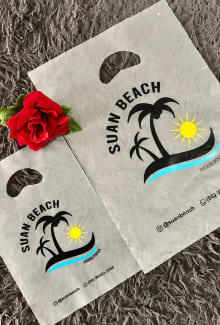 Sacola plástica alça vazada na cor branca, com a logomarca da Suan Beach (possui um desenho de duas palmeiras, na cor preto. As palmeiras estão encima de um pedaço de areia e ao redor tem água, simulando uma ilha. Ao fundo, tem um desenho de um sol).