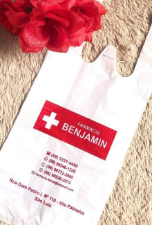 Sacola plástica camiseta na cor branca, com a logomarca Benjamin (possui uma palavra "Benjamin" e ao lado esquerdo dessa palavra tem um sinal de "+").